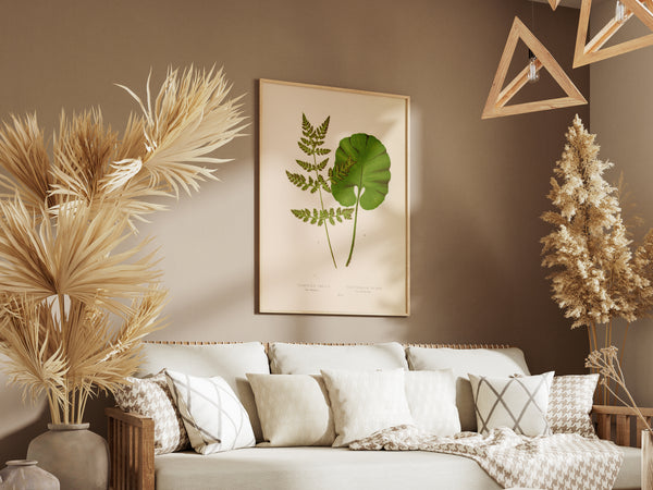 A framed botanical art print featuring green leaves on a cream background, displayed on a warm brown wall above a white sofa with neutral-toned pillows and dried pampas grass decor in a styled living room.