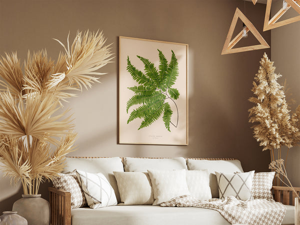 A framed botanical art print of a green fern leaf hanging on a warm brown wall above a beige sofa with patterned pillows and a throw blanket, styled with dried pampas grass in a cozy living room setting.